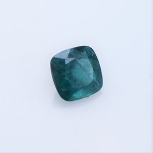 May include: A cushion-cut gemstone with a deep teal-green color and a faceted surface. The stone displays internal clarity and light reflection across its polished top, showcasing a square shape with rounded corners and a translucent appearance.