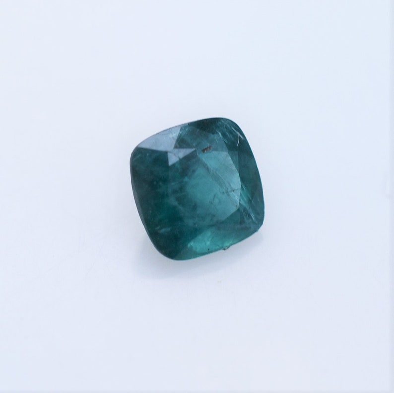 May include: A cushion-cut gemstone with a deep teal-green color and a faceted surface. The stone displays internal clarity and light reflection across its polished top, showcasing a square shape with rounded corners and a translucent appearance.