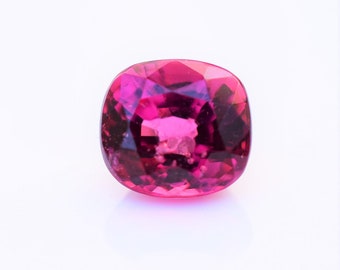 May include: A cushion-cut gemstone with a vibrant, deep magenta-pink colour. The stone features a faceted surface that reflects light, creating a brilliant appearance with visible internal clarity and sharp, geometric edges throughout the cut.