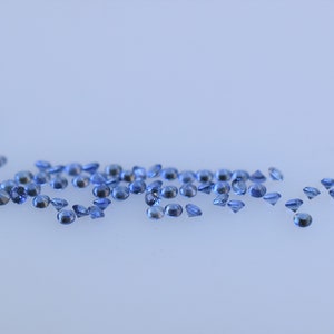 Op de afbeelding: Een verzameling kleine, ronde, gefacetteerde blauwe edelstenen verspreid over een effen, lichtgekleurd oppervlak. Elke individuele steen heeft een briljant slijpvorm met meerdere reflecterende vlakken die het licht vangen en blauwtinten tonen.