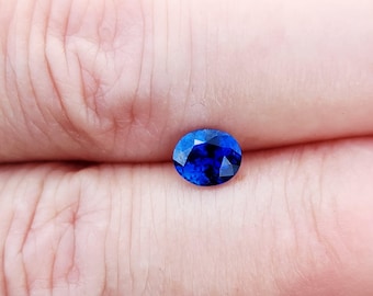 May include: A small, round, faceted gemstone with a deep, vibrant blue color. The stone features multiple triangular and kite-shaped facets that reflect light, creating a brilliant, sparkling appearance across its polished surface.