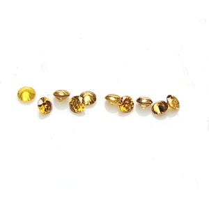 May include: A collection of nine small, round, faceted gemstones in varying shades of golden yellow and amber brown. Each stone features a brilliant cut with multiple reflective surfaces that catch the light, arranged in a loose horizontal line.