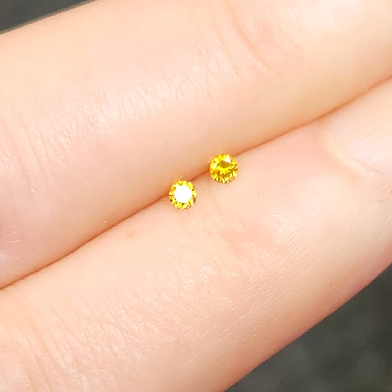 May include: Two small, round, brilliant-cut yellow gemstones with a faceted surface that reflects light. The stones are positioned side-by-side, displaying a vibrant, saturated golden-yellow hue and a circular shape with multiple sharp facets.
