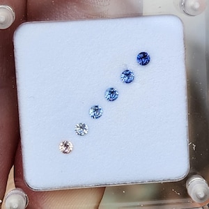 May include: A collection of round gemstones in varying shades of blue and one peach-colored stone, arranged in a diagonal line on a white surface. The gemstones are displayed in a clear plastic container, showcasing their color gradients.