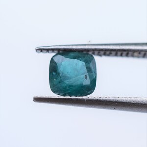 May include: A cushion-cut teal gemstone held by metal tweezers. The stone features a faceted surface with visible internal inclusions and a deep blue-green color, showcasing a translucent quality with light reflecting off the polished facets.