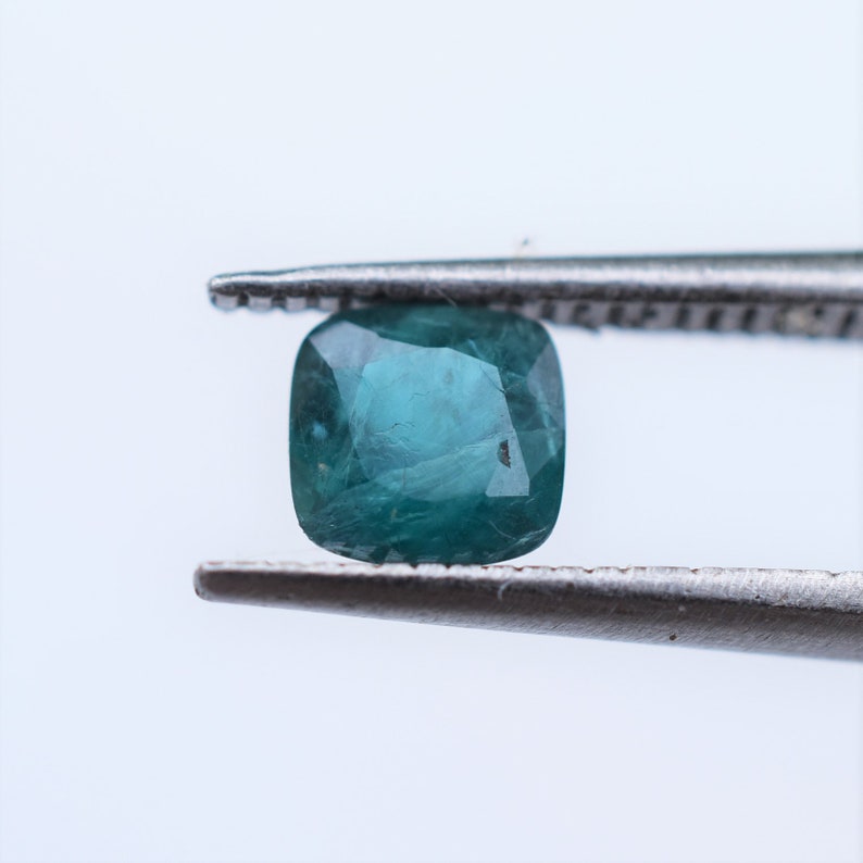 May include: A cushion-cut teal gemstone held by metal tweezers. The stone features a faceted surface with visible internal inclusions and a deep blue-green color, showcasing a translucent quality with light reflecting off the polished facets.