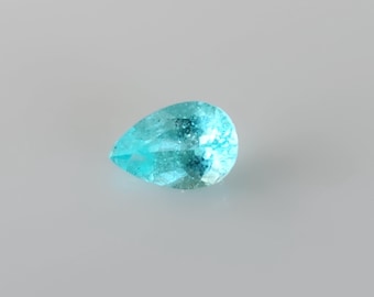 May include: A pear-shaped gemstone with a vibrant, translucent cyan-blue colour. The stone features a faceted surface that reflects light, creating a sparkling appearance. The gem is displayed against a neutral, light grey background.