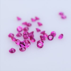 Pode incluir: Uma dispersão de pedras preciosas cor-de-rosa sobre uma superfície branca. As pedras preciosas são redondas e têm um acabamento brilhante e facetado.