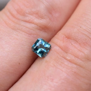 Op de afbeelding: Een vierkant geslepen edelsteen met een diepe blauwgroene kleur en een gefacetteerd oppervlak dat licht reflecteert. De steen heeft een helder, transparant uiterlijk met scherpe randen en een symmetrische vorm, met een levendige en verzadigde blauwgroene tint.