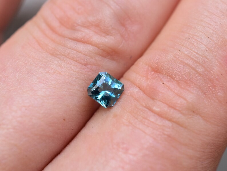 Op de afbeelding: Een vierkant geslepen edelsteen met een diepe blauwgroene kleur en een gefacetteerd oppervlak dat licht reflecteert. De steen heeft een helder, transparant uiterlijk met scherpe randen en een symmetrische vorm, met een levendige en verzadigde blauwgroene tint.