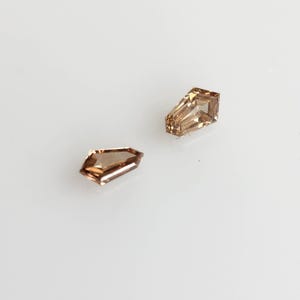May include: Two loose shield-cut gemstones with a light brown, champagne-colored hue. Each stone features a faceted surface that reflects light, displaying a geometric shape with a pointed end and a flat top edge, set against a plain white background.
