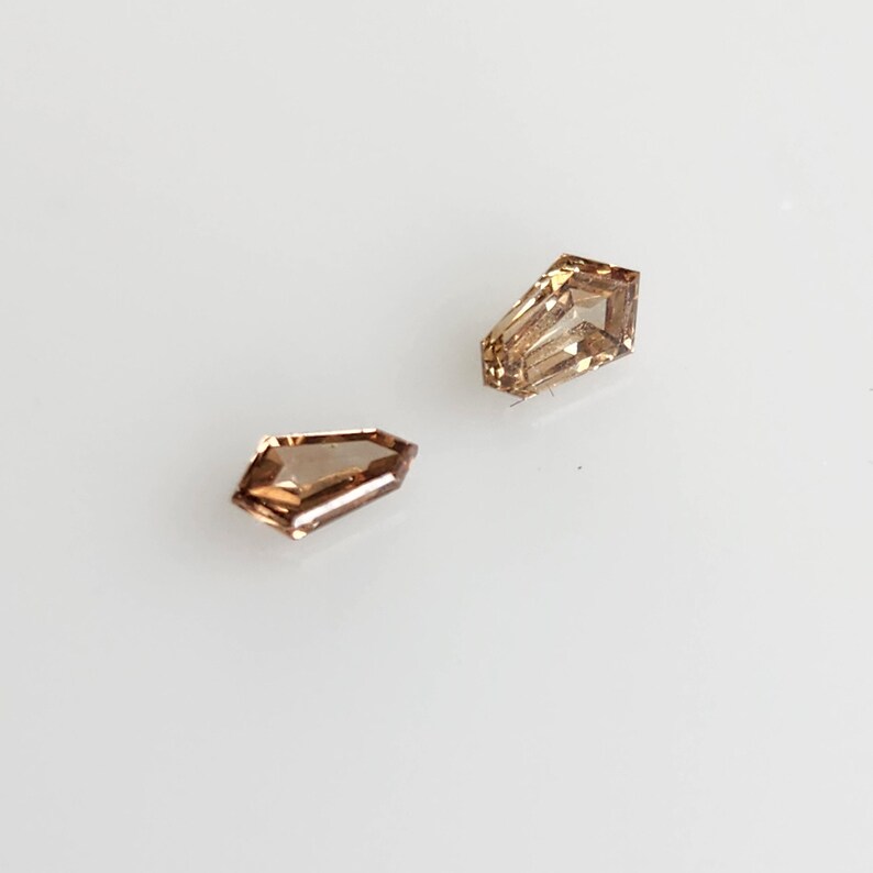 May include: Two loose shield-cut gemstones with a light brown, champagne-colored hue. Each stone features a faceted surface that reflects light, displaying a geometric shape with a pointed end and a flat top edge, set against a plain white background.