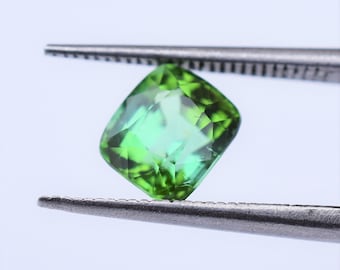 May include: A cushion-cut loose gemstone with a vibrant, translucent green color and multiple facets that reflect light. The gemstone is held securely between the serrated metal tips of a pair of silver-toned tweezers against a plain background.