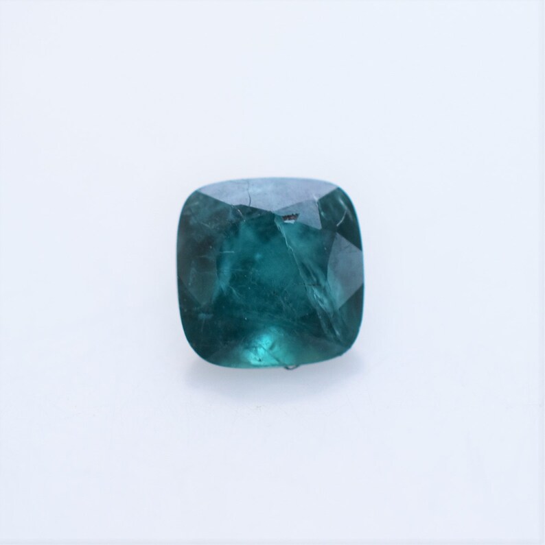 May include: A cushion-cut gemstone with a deep teal and blue-green color. The stone features a faceted surface that reflects light, showing internal inclusions and a translucent quality. The shape is square with rounded corners and smooth edges.