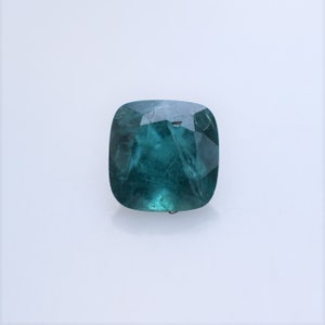 May include: A cushion-cut gemstone with a deep teal-green color and a faceted surface. The stone displays internal inclusions and a translucent quality, reflecting light across its polished, angular edges against a plain, light-colored background.