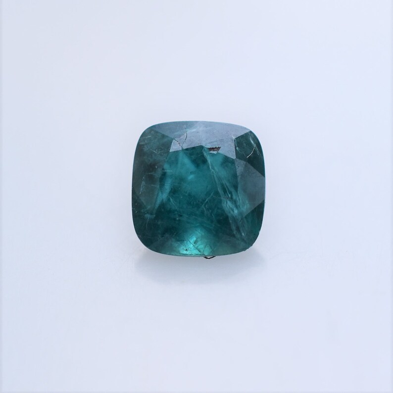 May include: A cushion-cut gemstone with a deep teal-green color and a faceted surface. The stone displays internal inclusions and a translucent quality, reflecting light across its polished, angular edges against a plain, light-colored background.