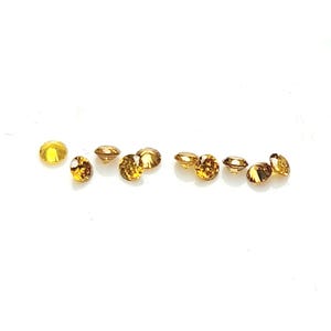 May include: A collection of nine small, round, faceted gemstones in a deep golden-brown hue. Each stone features a brilliant cut with multiple reflective surfaces that catch the light, arranged in a loose horizontal line against a plain background.