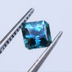 Op de afbeelding: Een vierkant geslepen edelsteen met een levendig blauw en groenblauw kleurenpalet, voorzien van meerdere facetten die het licht reflecteren. De edelsteen wordt vastgehouden door een zilverkleurige metalen pincet, wat de transparantie en geometrische vorm benadrukt.