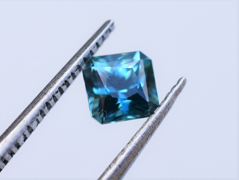 Op de afbeelding: Een vierkant geslepen edelsteen met een levendig blauw en groenblauw kleurenpalet, voorzien van meerdere facetten die het licht reflecteren. De edelsteen wordt vastgehouden door een zilverkleurige metalen pincet, wat de transparantie en geometrische vorm benadrukt.
