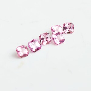 May include: Five loose, square-cut gemstones in a vibrant pink hue. Each stone features a faceted surface that reflects light, creating a brilliant, sparkling appearance. The gems are arranged in a slightly staggered, diagonal line formation.
