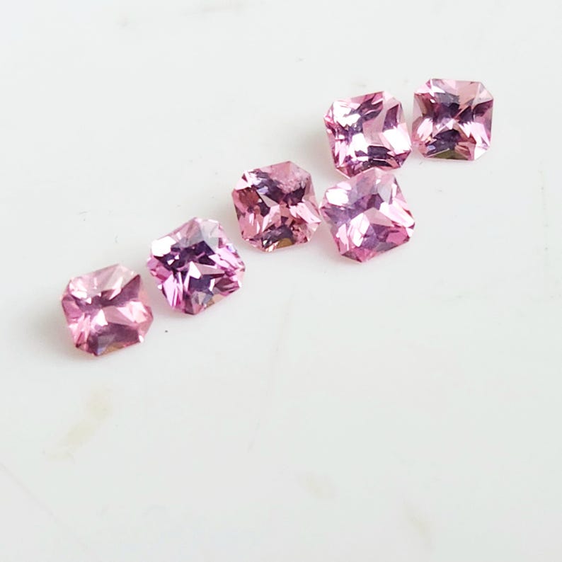 May include: Six loose, square-cut gemstones in a vibrant pink hue. Each stone features a faceted surface that reflects light, creating a sparkling appearance. The gems are arranged in a scattered, diagonal formation across the frame.