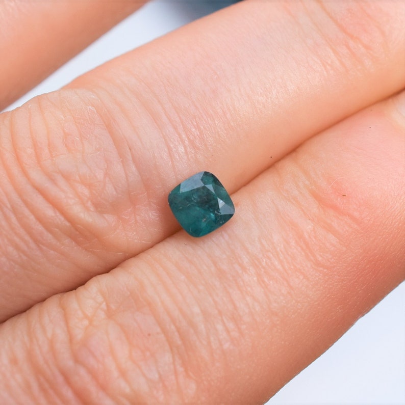 May include: A loose, cushion-cut gemstone with a deep teal-green color and faceted surface. The stone features a square shape with rounded corners, displaying internal clarity and light reflection across its polished, multi-faceted top surface.