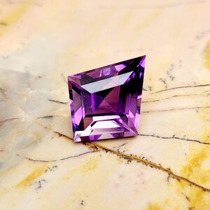 May include: A faceted purple amethyst gemstone with a kite-shaped silhouette. The stone features a geometric cut with sharp angles and a pointed top, displaying deep violet hues and light-reflecting surfaces throughout its transparent structure.