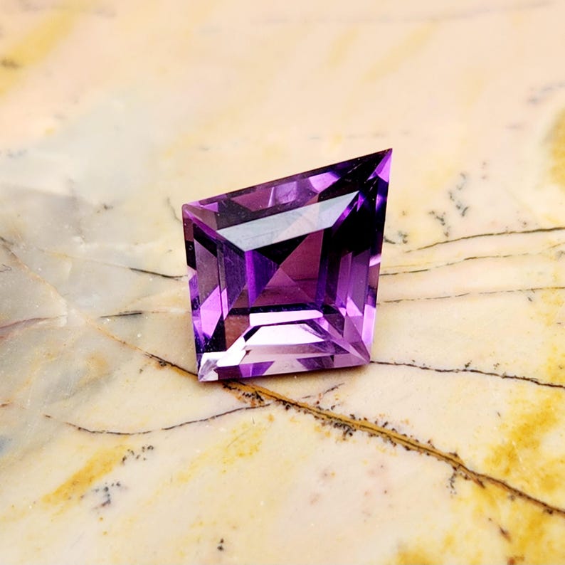 May include: A faceted purple amethyst gemstone with a kite-shaped silhouette. The stone features a geometric cut with sharp angles and a pointed top, displaying deep violet hues and light-reflecting surfaces throughout its transparent structure.