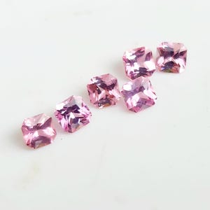 May include: Six loose, square-cut pink gemstones with faceted surfaces. Each stone displays a vibrant, translucent pink hue with sharp, geometric edges and a brilliant, reflective quality that catches the light, arranged in a diagonal pattern.