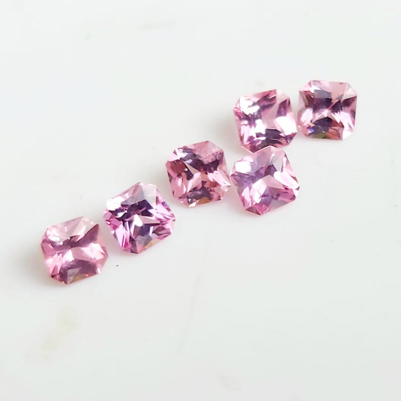 May include: Six loose, square-cut pink gemstones with faceted surfaces. Each stone displays a vibrant, translucent pink hue with sharp, geometric edges and a brilliant, reflective quality that catches the light, arranged in a diagonal pattern.