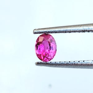 Puede incluir: Una gema ovalada de color rosa vibrante sostenida por unas pinzas.