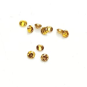 May include: A collection of eight small, round, faceted gemstones with a deep amber and golden-brown hue. Each stone features a brilliant cut with multiple reflective surfaces that catch the light, displaying a warm, translucent, and sparkling appearance.