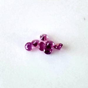 May include: Several faceted, oval-cut, deep pink gemstones are scattered on a white surface. The gemstones vary in size and cut, displaying a brilliant, reflective quality, indicating high clarity and polish. The presentation is clean and elegant, showcasing the gems' beauty.