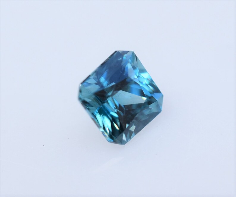 Op de afbeelding: Een vierkant geslepen edelsteen met een levendige blauwe tint en een gefacetteerd oppervlak. De steen vertoont diep marineblauwe en heldere cyaan tinten in de transparante structuur en reflecteert het licht met scherpe randen.