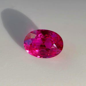 May include: A vibrant, oval-cut gemstone in a deep, saturated magenta-pink hue. The stone features a faceted surface that reflects light, creating internal patterns and highlights across its polished, translucent, and brilliant crystalline structure.