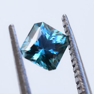 Op de afbeelding: Een vierkant geslepen edelsteen met facetten in een diepe blauwe en petrolkleur. De steen vertoont interne lichtreflecties en scherpe randen, vastgehouden door de gekartelde metalen punten van een pincet tegen een effen witte achtergrond.