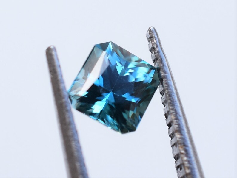 Op de afbeelding: Een vierkant geslepen edelsteen met facetten in een diepe blauwe en petrolkleur. De steen vertoont interne lichtreflecties en scherpe randen, vastgehouden door de gekartelde metalen punten van een pincet tegen een effen witte achtergrond.