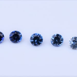 May include: Five round faceted gemstones arranged in a horizontal row. The stones display a color gradient, starting with two deep dark blue stones on the left, followed by two medium blue stones, and ending with one light pale blue stone on the right.
