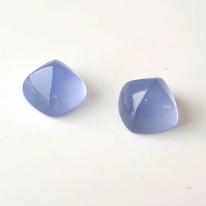 K&ouml;nnte beinhalten: Zwei polierte, hellblaue Edelsteine mit quadratischer Form und abgerundeten Kanten. Die Steine haben eine gl&auml;nzende Oberfl&auml;che und reflektieren das Licht, vor wei&szlig;em Hintergrund. Geeignet f&uuml;r Schmuckherstellung.