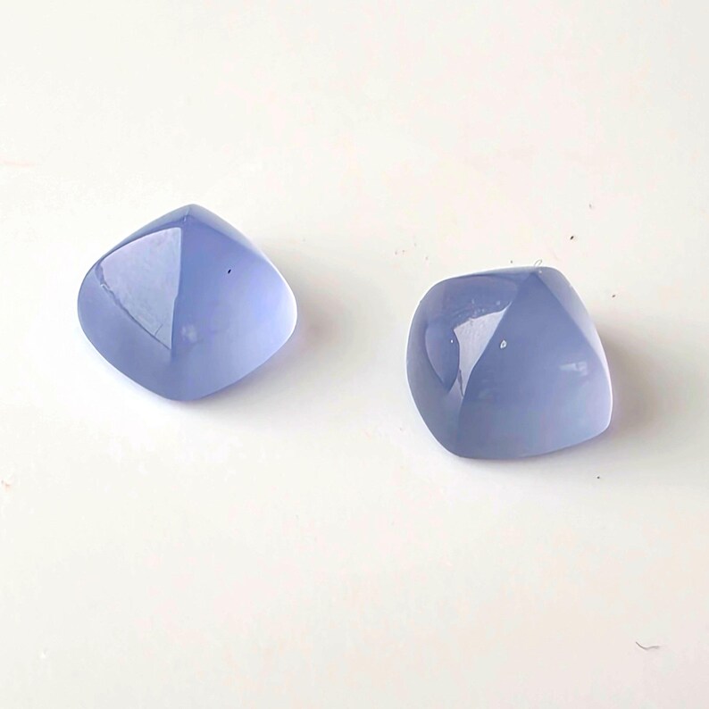 K&ouml;nnte beinhalten: Zwei polierte, hellblaue Edelsteine mit quadratischer Form und abgerundeten Kanten. Die Steine haben eine gl&auml;nzende Oberfl&auml;che und reflektieren das Licht, vor wei&szlig;em Hintergrund. Geeignet f&uuml;r Schmuckherstellung.
