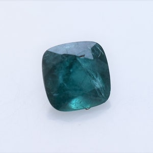 May include: A cushion-cut gemstone with a deep teal-green color and a faceted surface. The stone displays internal clarity characteristics and light reflections across its polished top, showcasing a square shape with rounded corners.