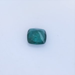May include: A loose, cushion-cut gemstone with a deep teal-green color. The stone features a faceted surface that reflects light, showcasing a translucent quality and a square shape with rounded corners, presented against a plain white background.