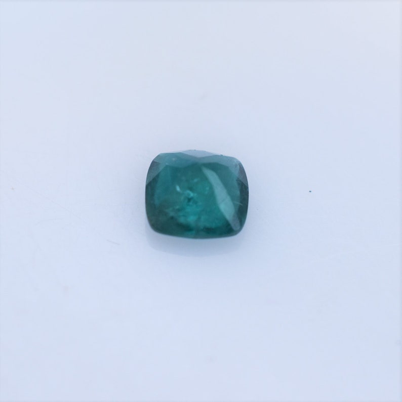 May include: A loose, cushion-cut gemstone with a deep teal-green color. The stone features a faceted surface that reflects light, showcasing a translucent quality and a square shape with rounded corners, presented against a plain white background.