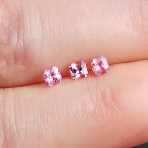 May include: Three small, square-cut pink gemstones with faceted surfaces that reflect light. The stones are arranged in a horizontal row, each displaying a vibrant, translucent pink hue and a precise geometric shape with sharp, clean edges.