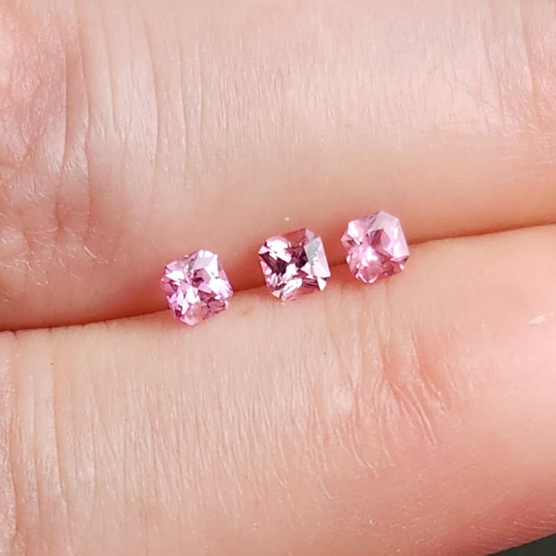 May include: Three small, square-cut pink gemstones with faceted surfaces that reflect light. The stones are arranged in a horizontal row, each displaying a vibrant, translucent pink hue and a precise geometric shape with sharp, clean edges.
