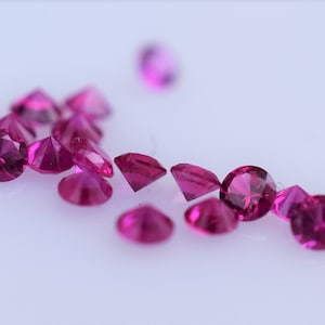 May include: A collection of loose pink gemstones, likely rubies, scattered on a white surface. The gemstones are round and faceted, with a variety of sizes.