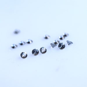 Puede incluir: Un grupo de pequeñas piedras preciosas redondas, transparentes, con un ligero tono azul. Las piedras preciosas están dispersas sobre una superficie blanca.