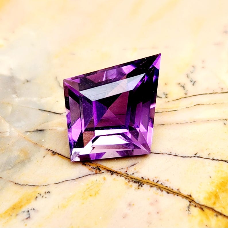 May include: A faceted purple gemstone with a kite-like geometric shape. The stone features sharp, angular edges and a brilliant, transparent violet hue with deep internal reflections and a polished surface that catches the light.