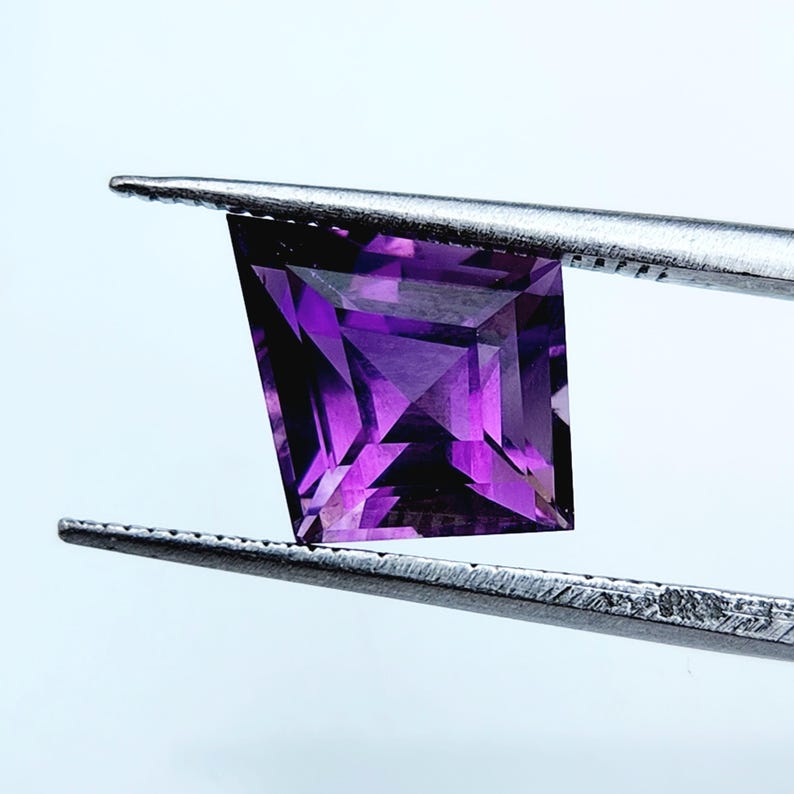 May include: A faceted purple gemstone with a square shape held by silver-toned metal tweezers. The gemstone displays deep violet hues and sharp geometric edges, reflecting light through its transparent, multi-faceted surface structure.