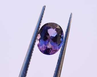 May include: A purple oval-shaped gemstone held by tweezers. The gemstone has a deep purple hue with a slight blue tint.